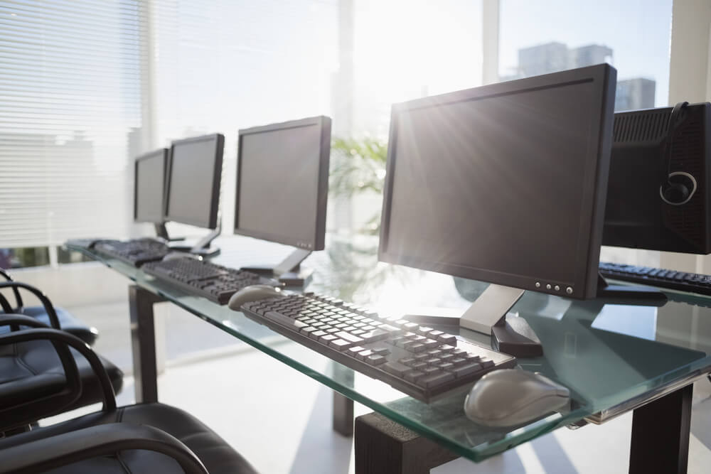 Several desktop computers on a glass desk with sun shining behind them