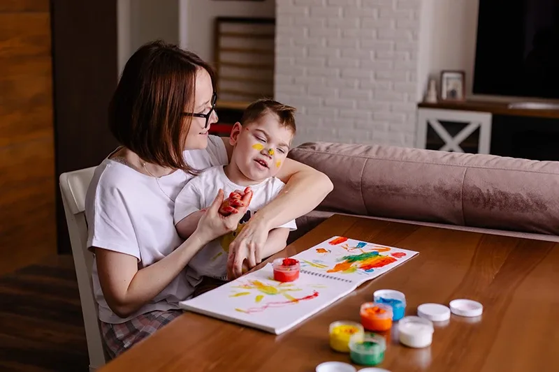 Woman holds toddler and teaches him to use finger paints on a notebook