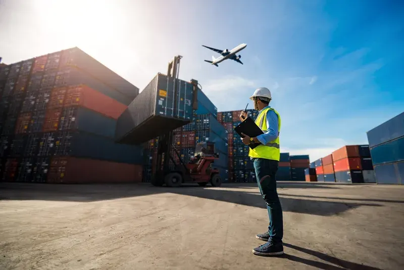 Man in yellow vest and hard hat directs another worker with heavy machinery to lift a shipping container into place while a plane flies overhead