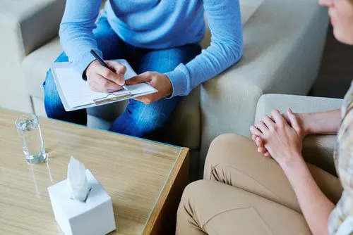 Therapist taking notes on a clipboard while speaking with a seated client during a counseling session.