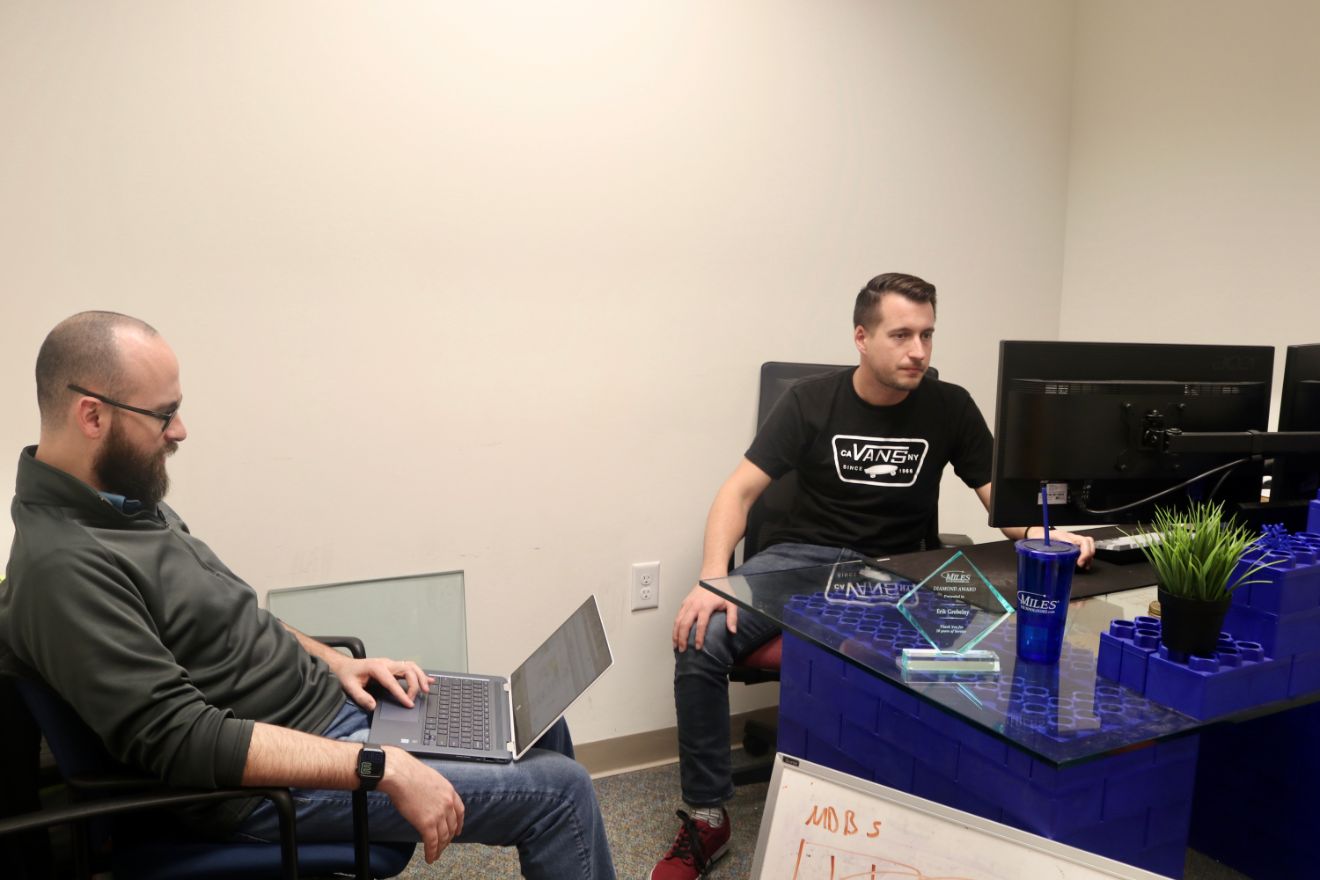 Two men in an office with a desk made of blocks, collaborating on software development using a desktop and laptop