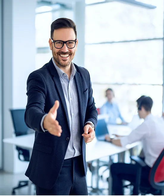 businessman welcoming new employee to his business team and company, giving hand forward in modern office with colleagues in the background.