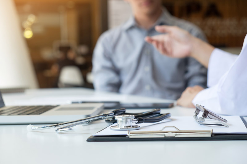 A desk with a laptop next to a clipboard, glasses, and stethoscope. There is a doctor and patient sitting next to the desk talking.