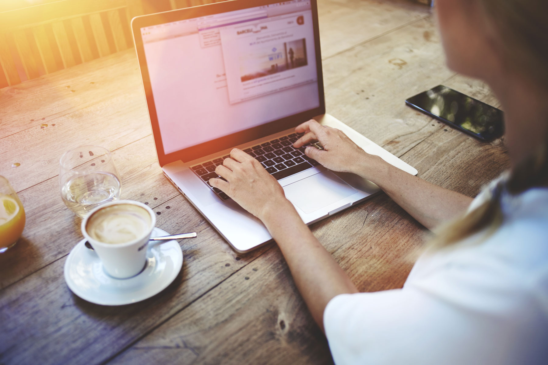 Woman sitting at a wooden table, looking at a page on the internet on a laptop with a cup of tea or coffee