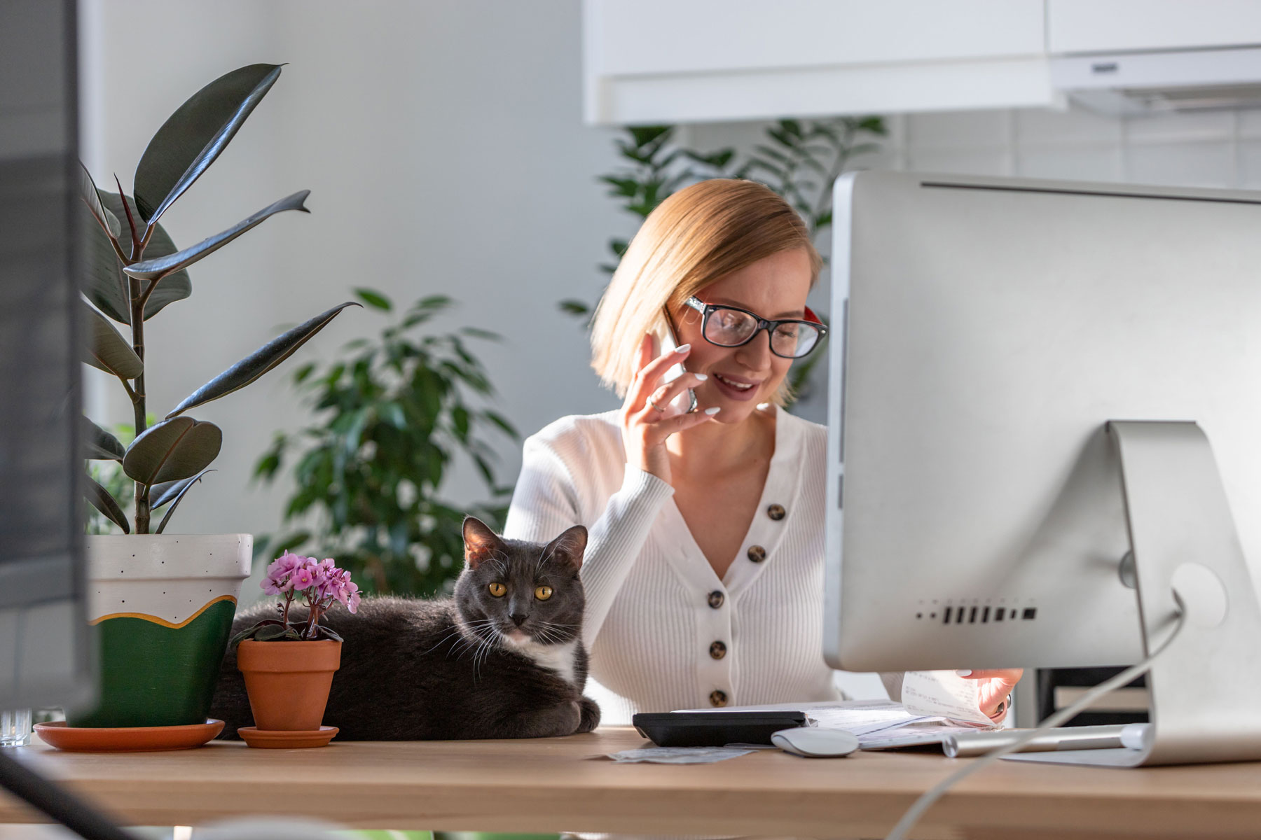 woman talking on the phone in front of a desktop computer monitor with a cat on the desk